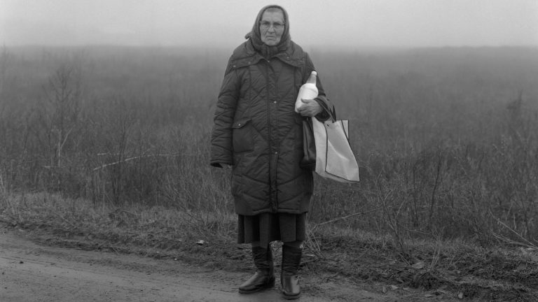 una donna anziana con una bottiglia di latte e una busta vuota della spesa posa frontale su una strada di campagna brulla