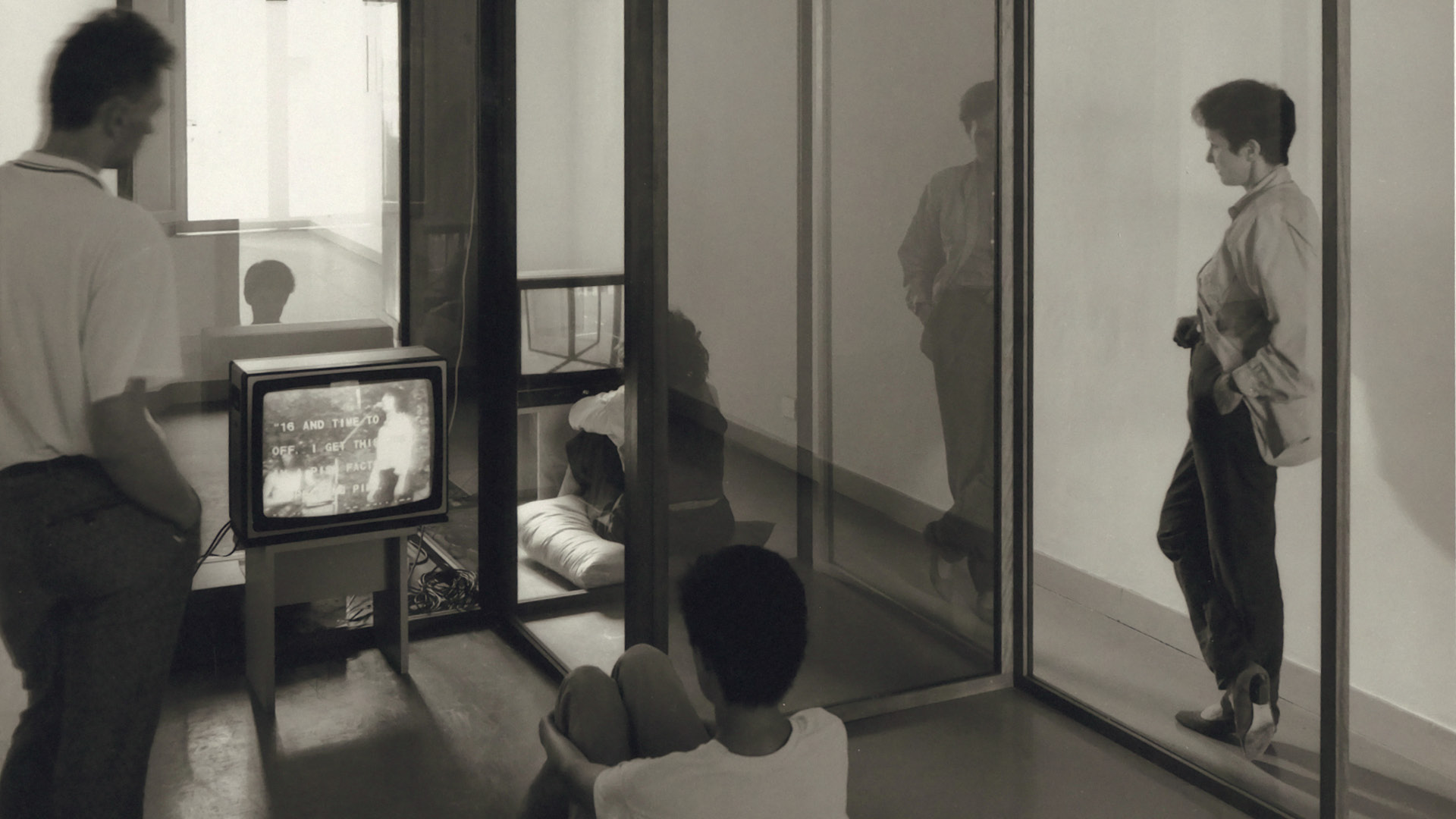 Fotografia in bianco e nero di un'installazione artistica in una galleria d'arte. Alcune persone osservano uno schermo televisivo che trasmette immagini in bianco e nero con scritte. Gli spettatori sono distribuiti nello spazio, tra vetri trasparenti e pareti minimali.