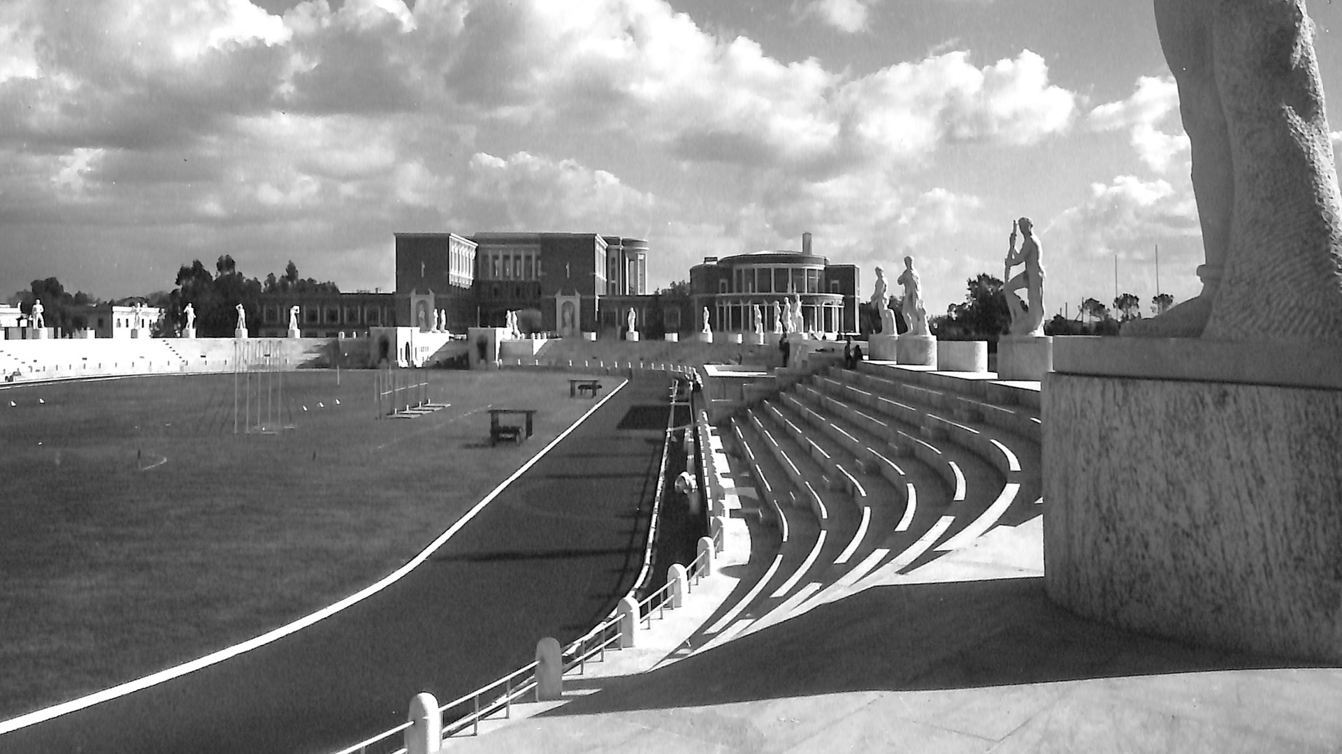 Veduta in bianco e nero dello Stadio dei Marmi al Foro Italico di Roma. In primo piano, gradinate curve in marmo e statue di atleti posizionate su piedistalli lungo il perimetro dello stadio. Sullo sfondo, edifici monumentali di architettura razionalista e un cielo con nuvole bianche.