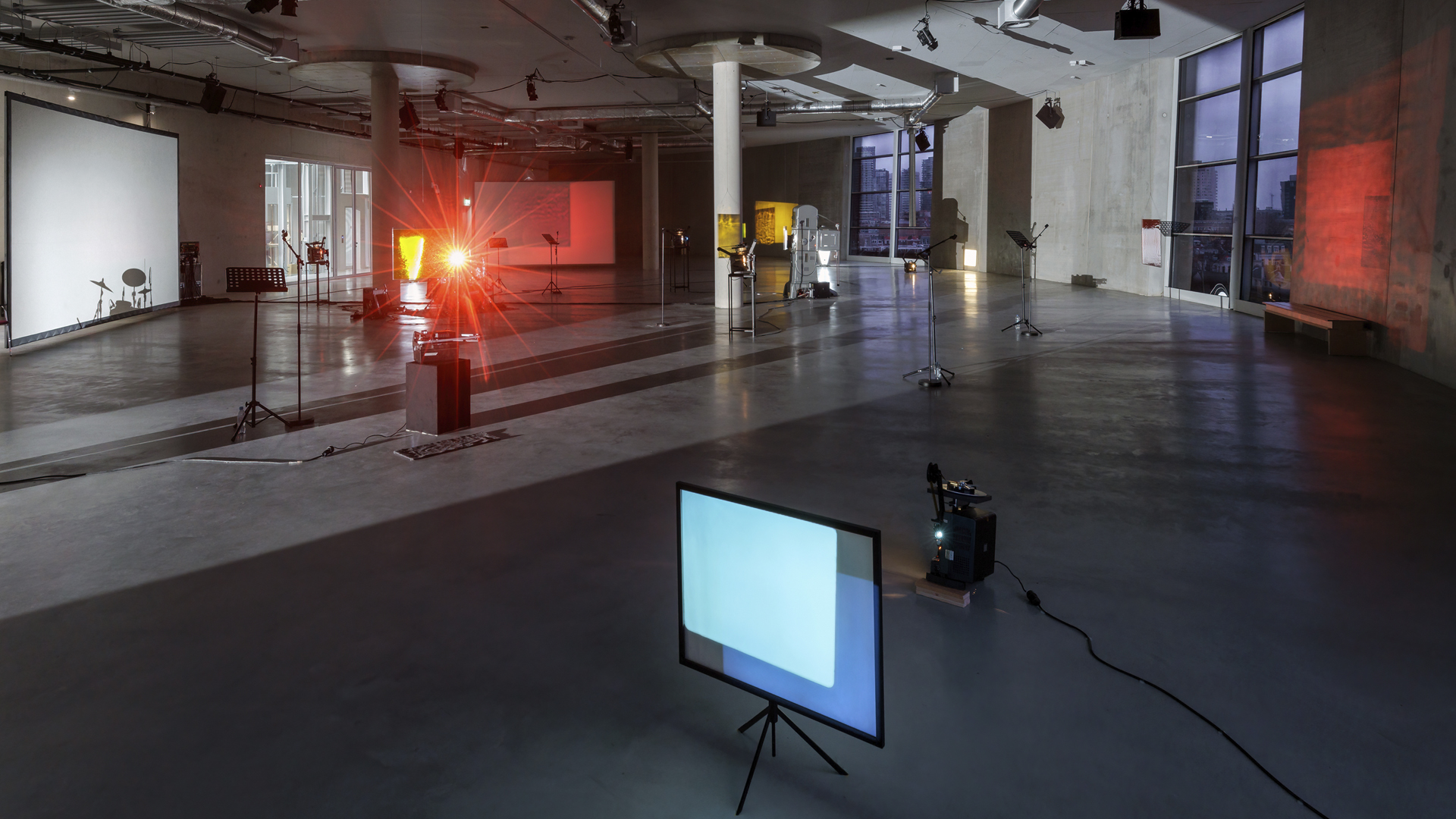 Spazio espositivo contemporaneo con pavimento in cemento levigato e ampie finestre. L'installazione include proiettori, schermi, leggii e strumenti musicali. La scena è illuminata da luci rosse e bianche che proiettano ombre e riflessi sulle pareti e sugli schermi.