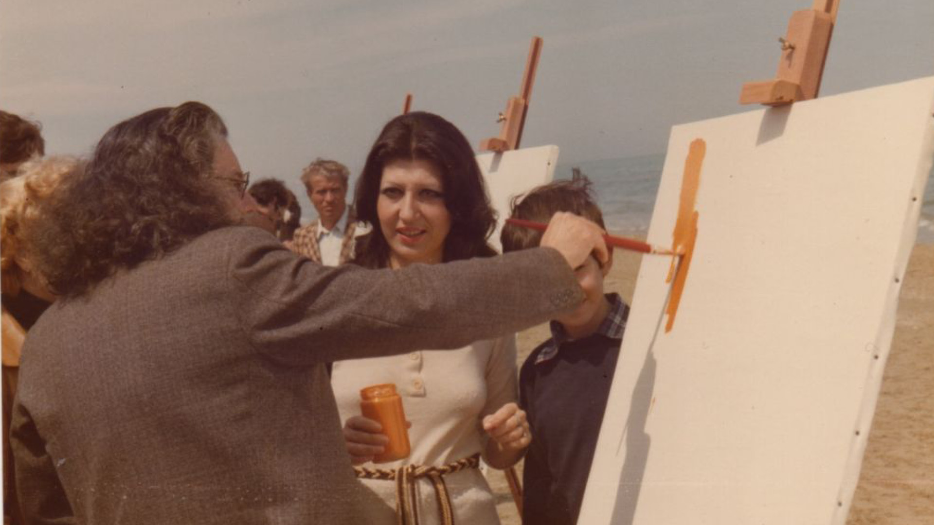 Fotografia a colori che ritrae un artista mentre dipinge su una tela all'aperto, in spiaggia. L'uomo con capelli lunghi e ricci tiene un pennello intinto nella vernice arancione, mentre una donna e un bambino osservano. Sullo sfondo, altre persone e il mare
