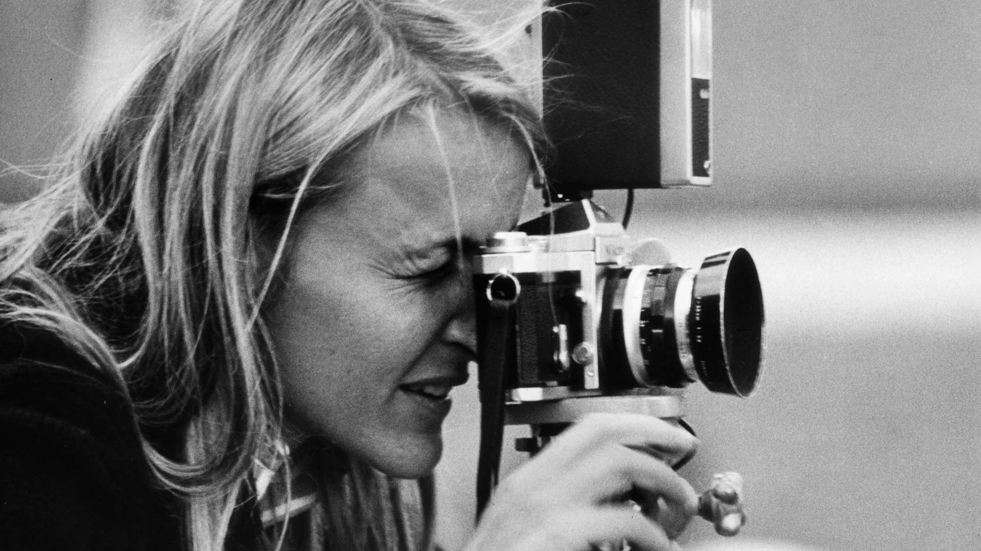 Primo piano in bianco e nero di una fotografa bionda mentre scatta una foto con una macchina fotografica analogica montata su treppiede. La donna è concentrata, con un occhio chiuso e la mano che regola l'obiettivo.