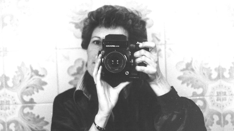 Autoritratto allo specchio in bianco e nero di Nanda Lanfranco, capelli ricci raccolti, che fotografa il proprio riflesso con una macchina fotografica biottica Bronica tenuta all'altezza del viso, coprendo gran parte del volto. Indossa un abito scuro. Sullo sfondo, piastrelle decorate a motivi floreali.
