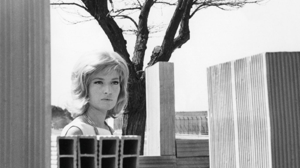 Fotogramma in bianco e nero: una donna (l'attrice Monica Vitti) con capelli biondi voluminosi e collana a più fili guarda leggermente di lato con espressione seria, in mezzo tra materiali edili accatastati. Sullo sfondo un albero spoglio e alcuni pannelli in legno e lamiera ondulata; in primo piano, sulla sinistra, blocchi di cemento forati. Ambientazione da cantiere o periferia urbana.