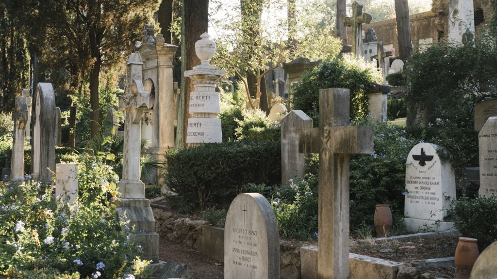Vista di un cimitero monumentale immerso nel verde, con lapidi e monumenti funebri di epoche diverse disposti tra alberi, cespugli fioriti di blu e vasi in terracotta. In primo piano una croce in pietra e alcune lapidi con iscrizioni leggibili, tra cui nomi italiani e un'iscrizione in inglese con simbolo di un uccello. Sullo sfondo, tra la vegetazione, si intravedono colonne, un'urna scolpita e un antico muro. Luce calda di fine giornata che filtra tra gli alberi. Fotografia a colori.