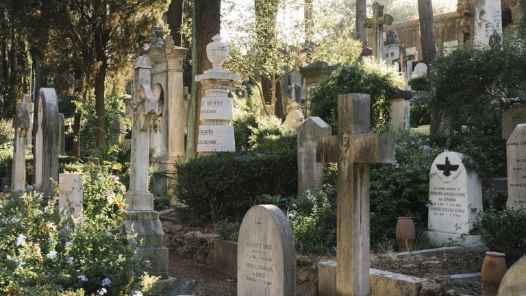 Vista di un cimitero monumentale immerso nel verde, con lapidi e monumenti funebri di epoche diverse disposti tra alberi, cespugli fioriti di blu e vasi in terracotta. In primo piano una croce in pietra e alcune lapidi con iscrizioni leggibili, tra cui nomi italiani e un'iscrizione in inglese con simbolo di un uccello. Sullo sfondo, tra la vegetazione, si intravedono colonne, un'urna scolpita e un antico muro. Luce calda di fine giornata che filtra tra gli alberi. Fotografia a colori.