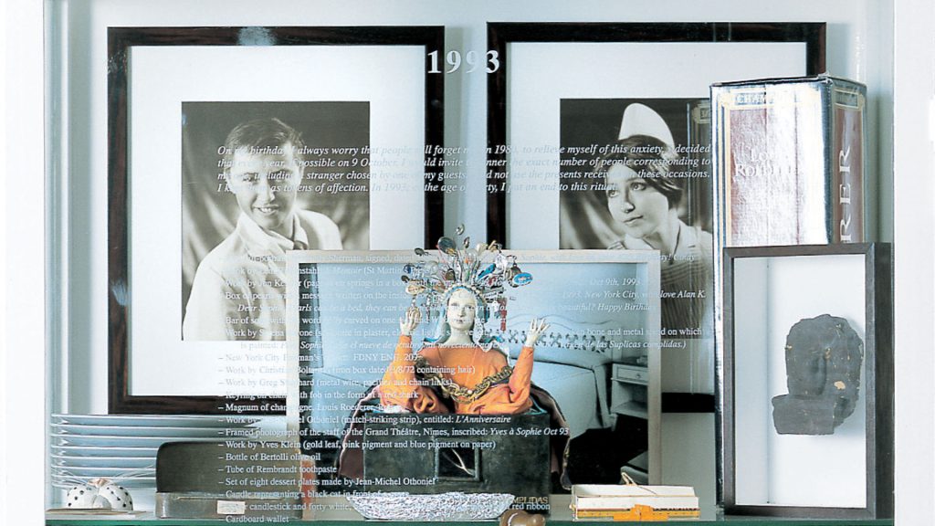 Installazione fotografica intitolata "1993", allestita come una vetrina-scaffale. Sopra, due grandi fotografie in bianco e nero incorniciate: a sinistra il ritratto di un giovane uomo, a destra quello di una donna con cuffia da infermiera; sovrapposto alle immagini, un testo in corsivo in cui l'autrice racconta di aver organizzato per anni, il giorno del proprio compleanno, una cena con tanti invitati quanti erano i suoi anni, conservando i regali ricevuti come pegni d'affetto, rituale poi interrotto nel 1993 al compimento dei quarant'anni. Sotto il testo, un lungo elenco elenca gli oggetti-regalo ricevuti in quell'occasione (tra cui opere di artisti, una scatola di perle, un pezzo di sapone, piatti da dessert, una candela a forma di gatto nero). In primo piano, al centro, una statuetta di figura femminile con copricapo piumato e abito arancione e dorato, le mani sollevate, posta su una base scura davanti a una cornice con una fotografia di un letto. Ai lati, altri oggetti esposti: un vassoio di piatti bianchi impilati, una scatola metallica con etichetta, una piccola scultura scura, una scatola di fiammiferi con etichetta "Chanel" sullo sfondo, e altre confezioni. Fotografia a colori.