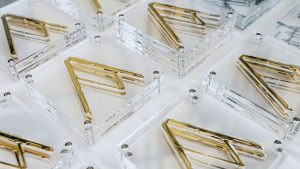 Vista dall'alto, in prospettiva diagonale, di una serie di targhe o premi identici disposti in file ordinate: basi trasparenti in plexiglass con angoli fissati da piccoli bulloni metallici, ciascuna con una lettera stilizzata in metallo dorato lucido al centro, simile a una "R" geometrica. Su alcune basi si intravedono incisioni con testo non leggibile. Fotografia a colori, a fuoco selettivo sugli esemplari in primo piano.