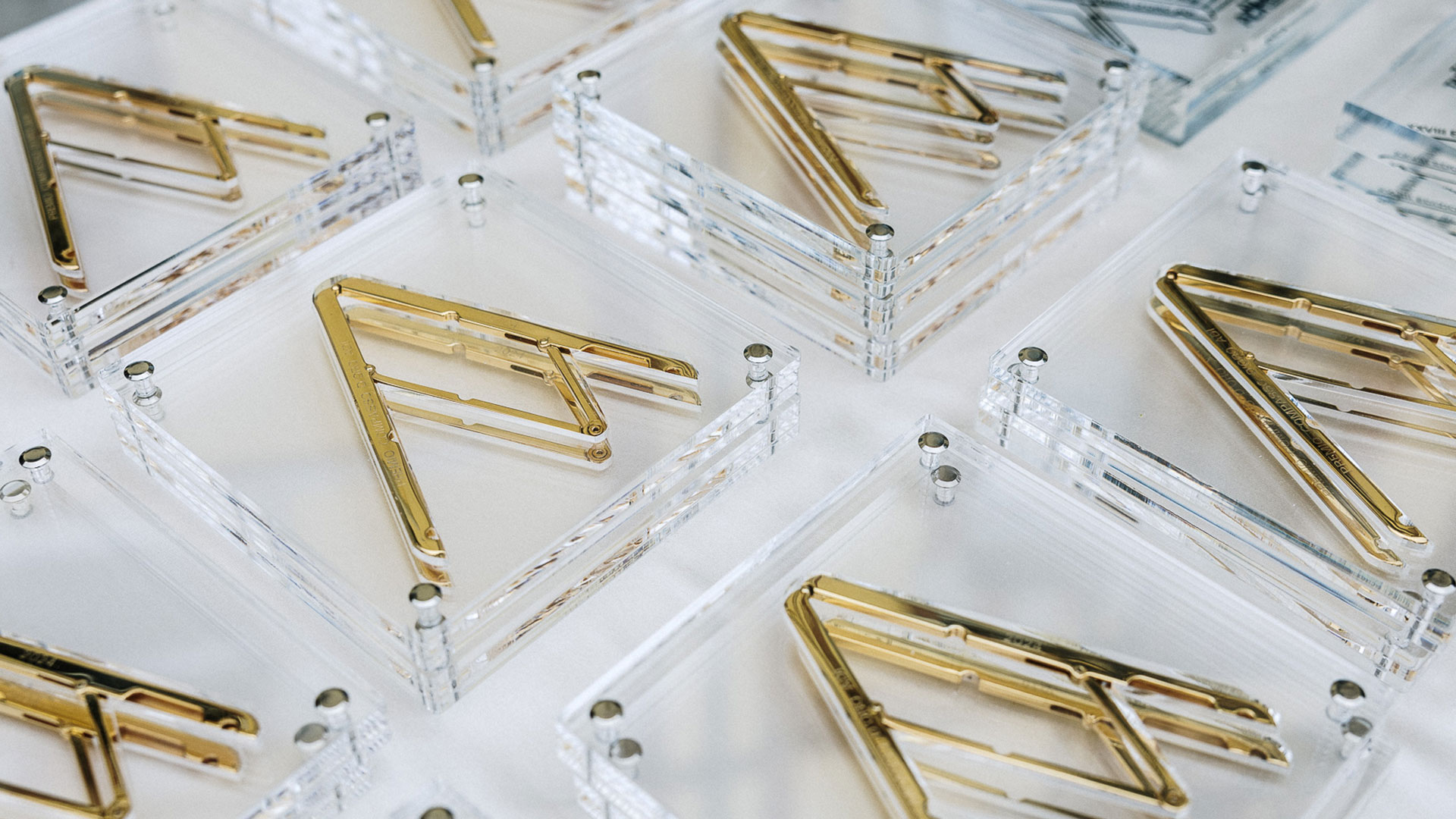 Vista dall'alto, in prospettiva diagonale, di una serie di targhe o premi identici disposti in file ordinate: basi trasparenti in plexiglass con angoli fissati da piccoli bulloni metallici, ciascuna con una lettera stilizzata in metallo dorato lucido al centro, simile a una "R" geometrica. Su alcune basi si intravedono incisioni con testo non leggibile. Fotografia a colori, a fuoco selettivo sugli esemplari in primo piano.