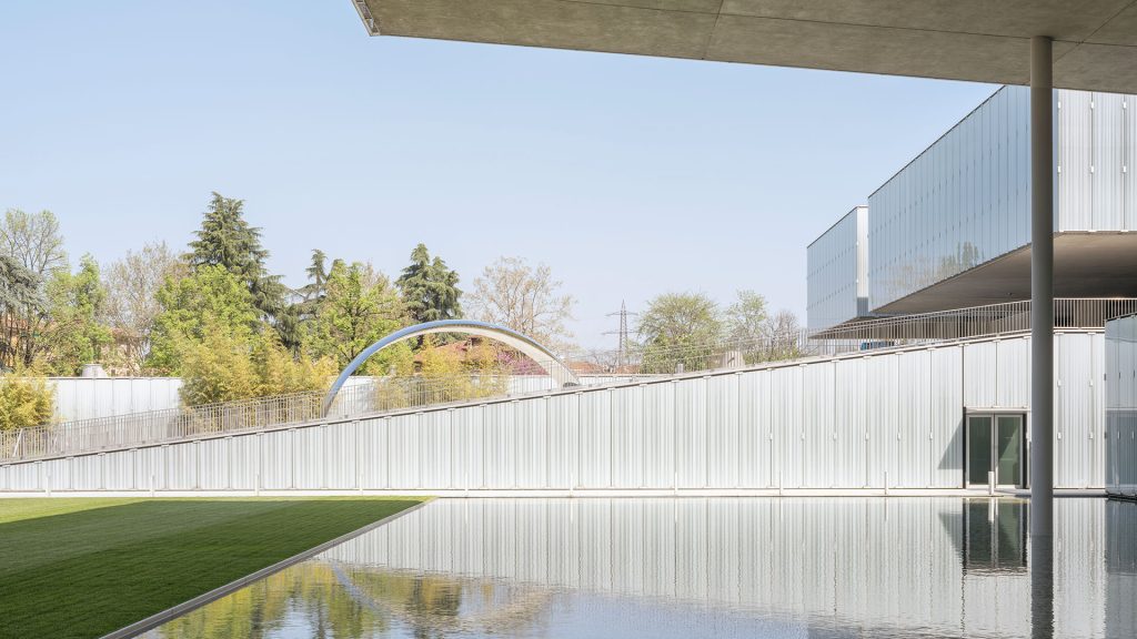 Scorcio architettonico di un edificio contemporaneo con rivestimento metallico verticale color argento, parzialmente coperto da una grande pensilina in cemento sorretta da un pilastro cilindrico. In primo piano, una vasca d’acqua a specchio affiancata da un prato verde, con un piccolo ponte ad arco in metallo sullo sfondo tra gli alberi. Cielo azzurro terso. Fotografia a colori.
