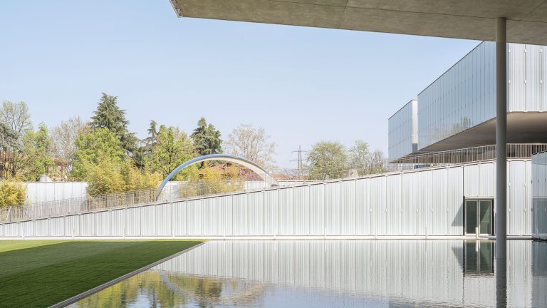 Scorcio architettonico di un edificio contemporaneo con rivestimento metallico verticale color argento, parzialmente coperto da una grande pensilina in cemento sorretta da un pilastro cilindrico. In primo piano, una vasca d’acqua a specchio affiancata da un prato verde, con un piccolo ponte ad arco in metallo sullo sfondo tra gli alberi. Cielo azzurro terso. Fotografia a colori.