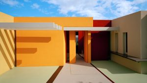 Facciata esterna di un edificio contemporaneo dai volumi squadrati e dalle pareti dipinte in colori accesi: giallo-arancio a sinistra, rosso e bordeaux al centro, beige a destra. Un corridoio aperto al centro, con pilastri arancioni e una pensilina bianca, lascia intravedere uno scorcio di mare sul fondo. La pavimentazione esterna è verde chiaro con bordi rossi. Cielo azzurro con nuvole sparse e ombre nette proiettate dal sole. Fotografia a colori.