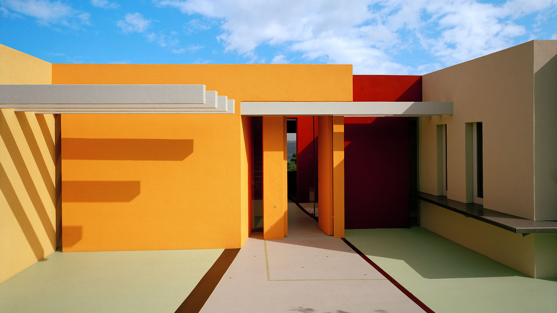 Facciata esterna di un edificio contemporaneo dai volumi squadrati e dalle pareti dipinte in colori accesi: giallo-arancio a sinistra, rosso e bordeaux al centro, beige a destra. Un corridoio aperto al centro, con pilastri arancioni e una pensilina bianca, lascia intravedere uno scorcio di mare sul fondo. La pavimentazione esterna è verde chiaro con bordi rossi. Cielo azzurro con nuvole sparse e ombre nette proiettate dal sole. Fotografia a colori.