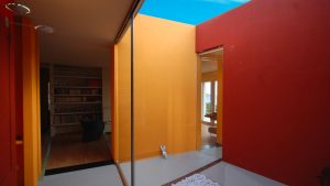 Interno di un’abitazione dal design contemporaneo con pareti dipinte in blocchi di colore vivace: giallo-arancio e rosso, con un’apertura nel soffitto da cui si vede il cielo azzurro. A sinistra, attraverso una porta, si intravede una stanza con libreria a parete e poltrona scura. A destra, un’altra apertura mostra una stanza luminosa con vista sul mare attraverso ampie vetrate. Sul pavimento in primo piano, una piccola vasca con ciottoli bianchi e una minuscola figura stilizzata bianca. Fotografia a colori.