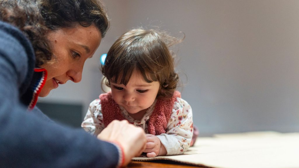 Primo piano di una donna china verso una bambina piccola, entrambe intente a osservare e toccare un foglio di cartone appoggiato su un tavolo.