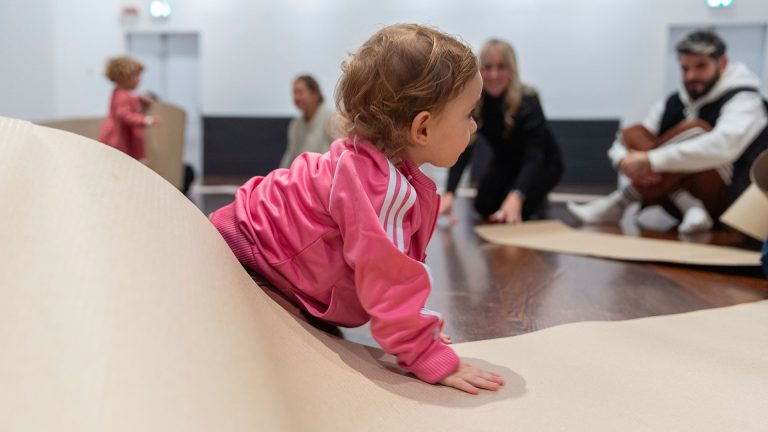 Una bambina in tuta rosa gattona su un foglio di cartone ondulato sollevato a formare un’onda. Sullo sfondo, altri bambini e adulti seduti a terra in una sala museale.
