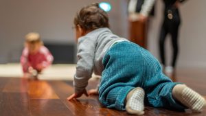 Un bambino visto di spalle gattona su un pavimento in legno, con un altro bambino sfocato sullo sfondo a sinistra e due figure in piedi sulla destra.