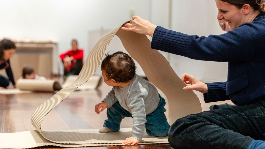 Una donna solleva un foglio di cartone ondulato a formare un arco, sotto il quale un bambino piccolo passa gattonando. Sullo sfondo, altri adulti e bambini seduti a terra tra fogli di cartone arrotolati, in una sala museale.