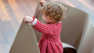Una bambina con capelli biondi ricci, vista di spalle, afferra con le mani il bordo di un grande foglio di cartone ondulato ripiegato a formare una struttura curva.