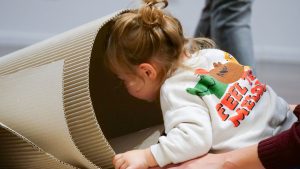 Primo piano di una bambina con i capelli raccolti che infila la testa dentro un tubo o cono formato da un foglio di cartone ondulato arrotolato, sorretta da un adulto.