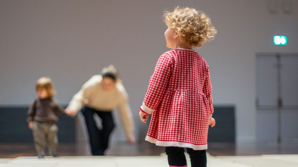 Una bambina con capelli ricci biondi, vista di spalle, osserva un adulto e un altro bambino sullo sfondo, fuori fuoco, in una sala museale con pavimento chiaro.