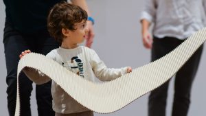 Un bambino con capelli ricci tiene con entrambe le mani un lungo foglio di cartone ondulato piegato a onda, guardando di lato verso un adulto fuori fuoco. Sullo sfondo, altre persone in una sala museale.