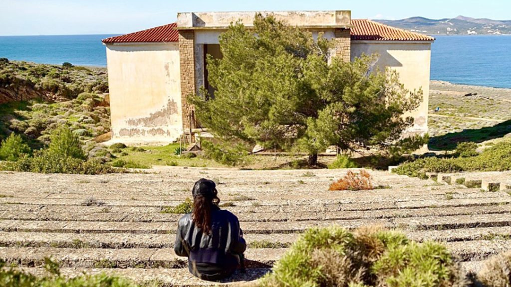 Una persona con berretto e capelli lunghi, vista di spalle, è seduta su una gradinata in pietra di fronte a un edificio abbandonato con tetto a coppi rossi e pareti intonacate scrostate, al centro del quale cresce un grande pino. Sullo sfondo, il mare e la costa con colline in lontananza; ai lati, vegetazione mediterranea bassa. Giornata di sole, cielo sereno. Fotografia a colori.