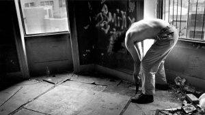 Fotografia in bianco e nero: un uomo a torso nudo, visto di profilo, è piegato in avanti in una stanza fatiscente con pareti scrostate e graffiti, intento a spaccare il pavimento con un attrezzo. Indossa pantaloni chiari abbassati sui fianchi. Sullo sfondo, finestre con inferriate affacciate su edifici urbani; a terra si intravedono una bottiglia vuota e detriti.