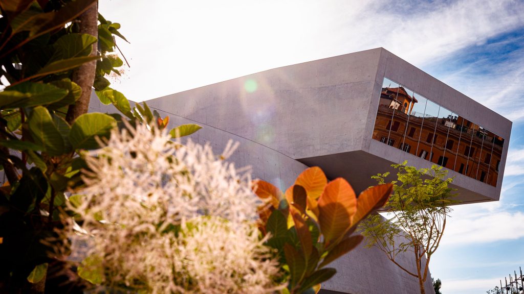 Dettaglio ravvicinato della vetrata del MAXXI che riflette un edificio color ruggine con persiane verdi, incorniciata da rami in primo piano con foglie verdi e rosse, molto sfocati. Cielo luminoso sullo sfondo. Fotografia a colori.