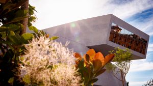 Dettaglio ravvicinato della vetrata del MAXXI che riflette un edificio color ruggine con persiane verdi, incorniciata da rami in primo piano con foglie verdi e rosse, molto sfocati. Cielo luminoso sullo sfondo. Fotografia a colori.