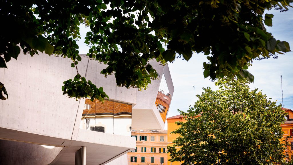 Dettaglio dell’edificio del MAXXI con la struttura in cemento a sbalzo e una vetrata che riflette un palazzo color ruggine, incorniciato da rami carichi di foglie verdi in primo piano, fuori fuoco. Cielo azzurro sullo sfondo. Fotografia a colori.