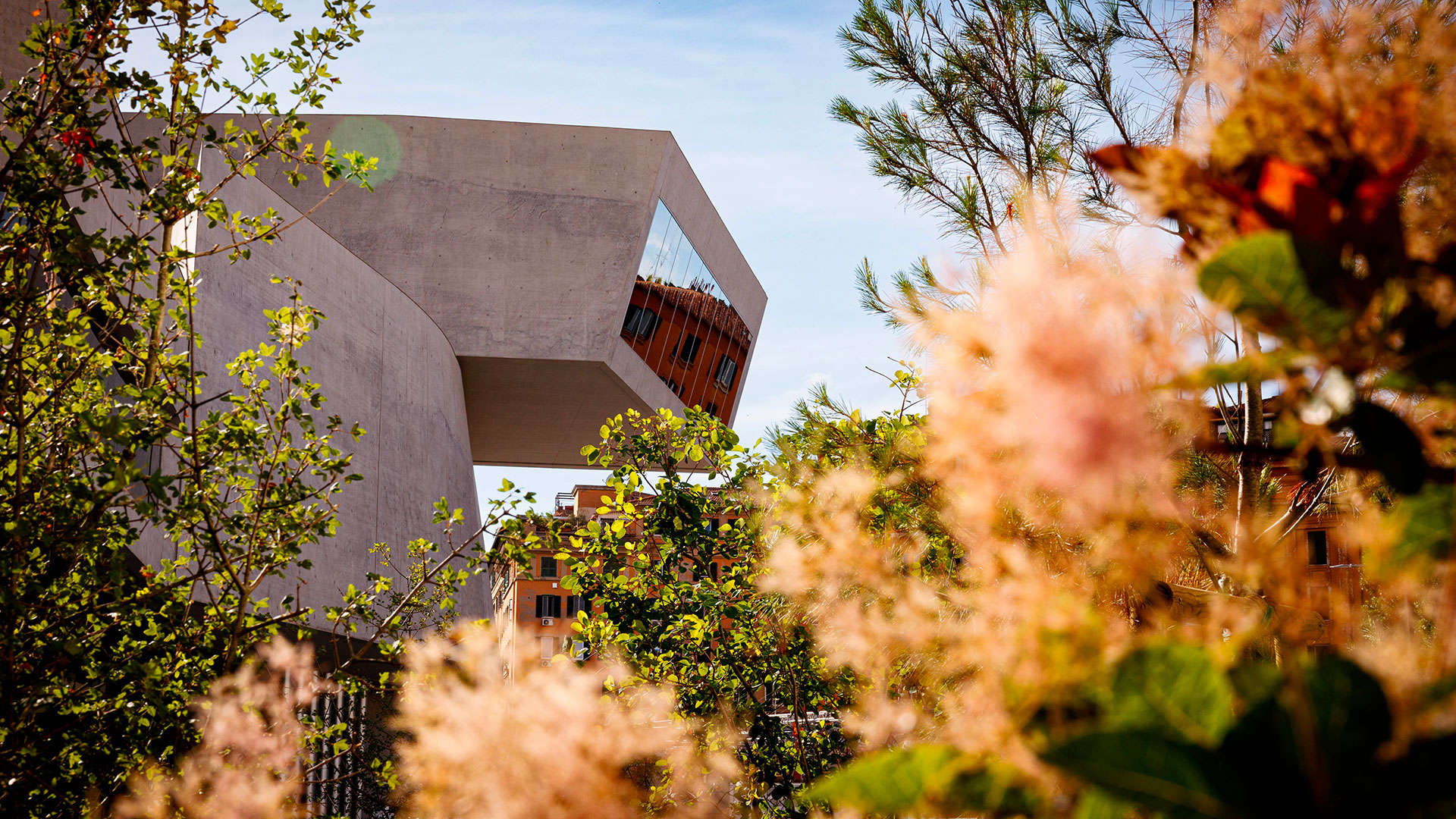 Vista dal basso della struttura in cemento del MAXXI, inquadrata tra foglie verdi e un ramo con infiorescenze rosa sfocate in primo piano. Sullo sfondo, un edificio color ruggine e cielo azzurro. Fotografia a colori.
