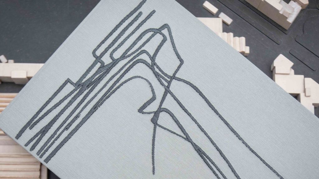Dettaglio ravvicinato di un libro con copertina in tessuto grigio chiaro, su cui è ricamato in filo grigio scuro un disegno architettonico astratto composto da linee curve e angolari intrecciate, simile alla pianta o al profilo di un edificio. Il libro è appoggiato in diagonale su un tavolo scuro, sullo sfondo del quale si intravede, sfocato, un plastico architettonico in legno chiaro. Fotografia a colori, in primo piano.