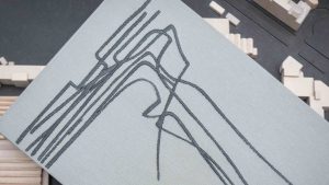Dettaglio ravvicinato di un libro con copertina in tessuto grigio chiaro, su cui è ricamato in filo grigio scuro un disegno architettonico astratto composto da linee curve e angolari intrecciate, simile alla pianta o al profilo di un edificio. Il libro è appoggiato in diagonale su un tavolo scuro, sullo sfondo del quale si intravede, sfocato, un plastico architettonico in legno chiaro. Fotografia a colori, in primo piano.