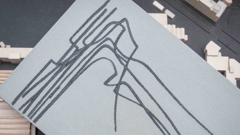 Dettaglio ravvicinato di un libro con copertina in tessuto grigio chiaro, su cui è ricamato in filo grigio scuro un disegno architettonico astratto composto da linee curve e angolari intrecciate, simile alla pianta o al profilo di un edificio. Il libro è appoggiato in diagonale su un tavolo scuro, sullo sfondo del quale si intravede, sfocato, un plastico architettonico in legno chiaro. Fotografia a colori, in primo piano.