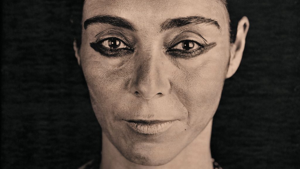 Ritratto fotografico in bianco e nero, fortemente ravvicinato, di una donna a capo rasato che guarda dritto verso l’obiettivo. Ha occhi scuri bordati da un marcato trucco nero allungato verso le tempie e sopracciglia folte e spesse. Lo sfondo è nero uniforme.
