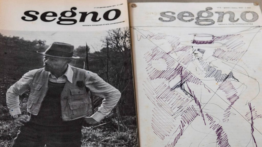 Due copertine affiancate della rivista "segno". A sinistra, una fotografia in bianco e nero di Joseph Beuys con cappello a tesa larga, giacca con molte tasche e mani sui fianchi, ritratto all'aperto tra vegetazione secca. A destra, un'altra copertina della stessa testata, su cui è sovrapposto un disegno a penna nei toni del viola e del nero: la figura schizzata di una persona con cappello, realizzata con tratti rapidi e tratteggi incrociati, in stile gestuale e astratto.
