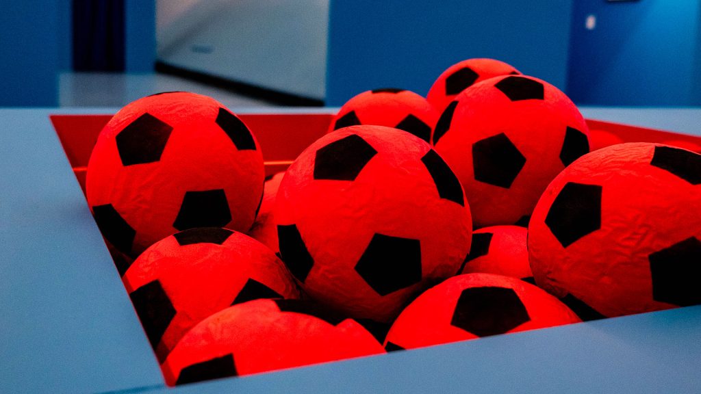 Primo piano di un gruppo di palloni da calcio rossi con disegno a pentagoni neri, ammassati dentro un contenitore triangolare rosso incassato in una struttura azzurra. Sullo sfondo, ambiente dai toni blu, fuori fuoco. Fotografia a colori.