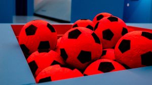 Primo piano di un gruppo di palloni da calcio rossi con disegno a pentagoni neri, ammassati dentro un contenitore triangolare rosso incassato in una struttura azzurra. Sullo sfondo, ambiente dai toni blu, fuori fuoco. Fotografia a colori.