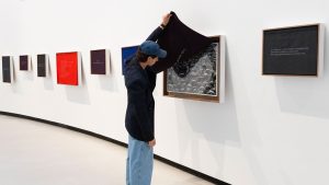 Un visitatore in cappellino e giacca scura solleva un panno di stoffa nera che copre un’opera incorniciata, rivelando una fotografia in bianco e nero di una folla vista dall’alto, con teste coperte di neve. Il lembo di stoffa sollevato reca una scritta ricamata capovolta, non leggibile nella foto. L’opera fa parte di un allestimento museale con una fila di cornici colorate (nere, viola, rosse, blu) contenenti testi, disposte lungo una parete bianca curva; una cornice più grande a destra mostra un testo in francese su fondo scuro. Fotografia a colori.